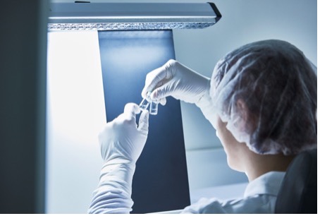 Woman inspecting two vials for particulates in front of a white and black background under bright lights.