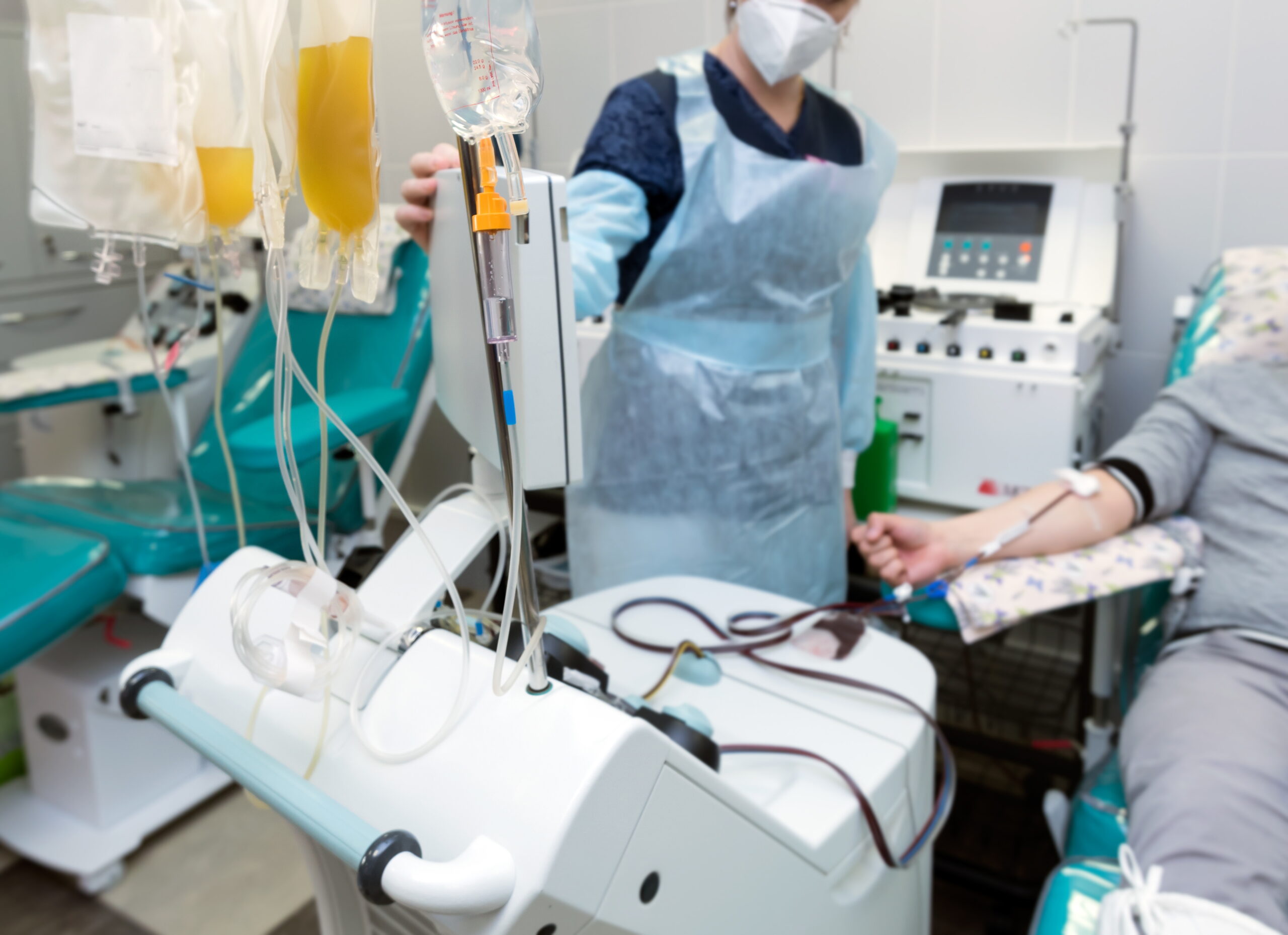 Patient undergoing apheresis procedure with blood drawn through tubing connected to a machine that separates blood components, while a healthcare worker in protective gear monitors the process.