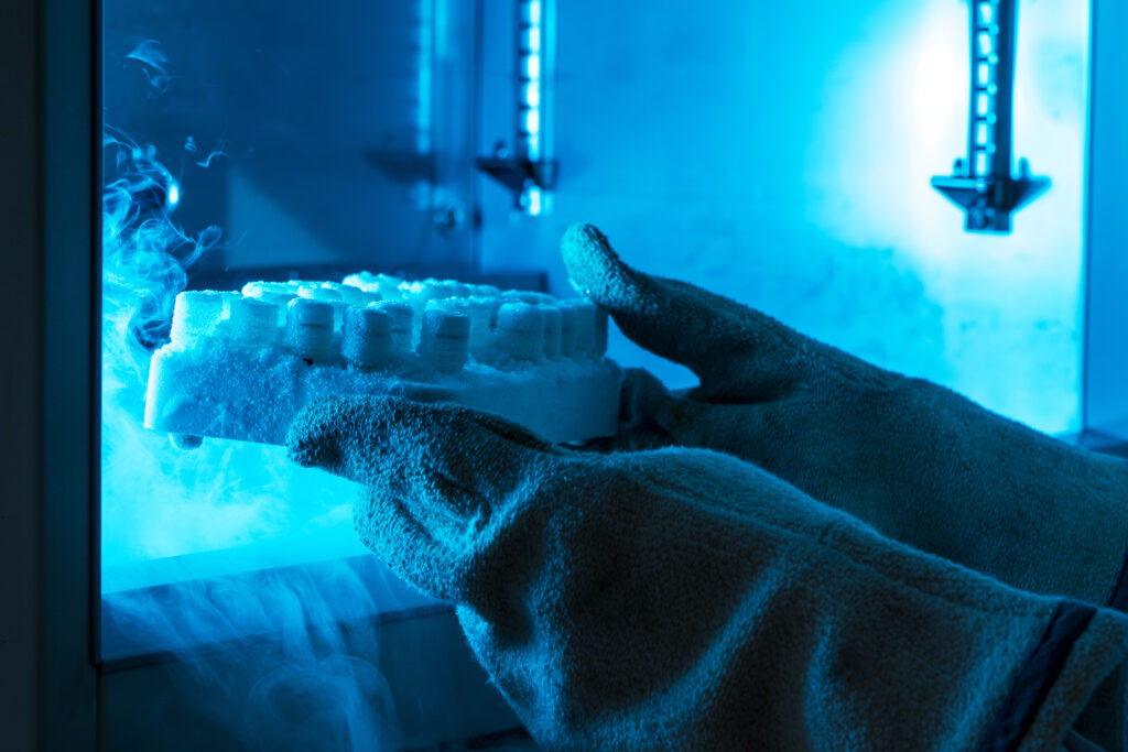 Close-up of insulated gloves holding a frosted rack of cryovials inside a blue-lit cryogenic freezer, with vapor rising from the frozen samples.